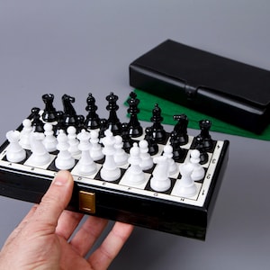 Puede incluir: Un juego de ajedrez magnético de viaje en blanco y negro con una caja negra. El tablero de ajedrez está abierto y las piezas están dispuestas para una partida. La caja está forrada con fieltro verde.