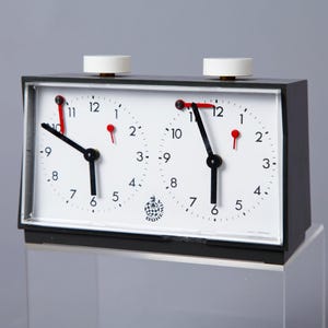 Può includere: Un orologio da scacchi nero con due quadranti bianchi. Ogni quadrante ha lancette nere e numeri neri. I quadranti sono impostati alle 12:00.