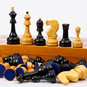 Op de afbeelding: Een houten schaakspel met zwarte en bruine stukken. De stukken zijn gerangschikt op een houten schaakbord, met enkele stukken rechtop en andere omver. De schaakstukken omvatten een koning, een dame, een loper, een paard, een toren en een pion.