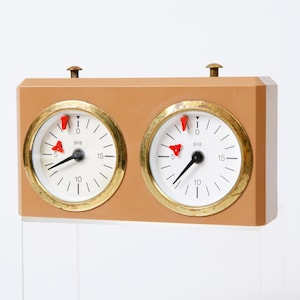 May include: A beige wooden chess clock with two round white faces. Each face has black hands and black numbers, and a red flag at the top.