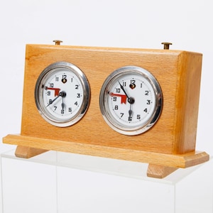 Op de afbeelding: Een houten schaakklok met twee ronde wijzerplaten. Elke wijzerplaat heeft een witte achtergrond met zwarte cijfers en wijzers, en een rode indicator. De klok is gemaakt van lichtbruin hout en heeft een eenvoudig, klassiek ontwerp.