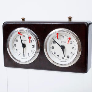 May include: A dark brown analog chess clock with two circular white faces, silver bezels, and black hands. Each face features the text BHB and Made in Germany. Two small brass-colored buttons are located on the top of the rectangular housing.