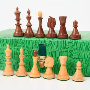 May include: A set of wooden chess pieces in a green leather case. The pieces are arranged in two rows, with the white pieces in the front and the black pieces in the back. The pieces are all carved in a traditional style.