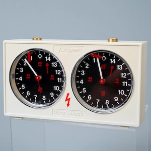 Può includere: Un orologio da scacchi analogico Jerger vintage con una custodia rettangolare color crema. Presenta due quadranti circolari neri con numeri bianchi, indicatori rossi e un simbolo di fulmine rosso tra di essi. Il testo riporta Jerger e blitz chess.