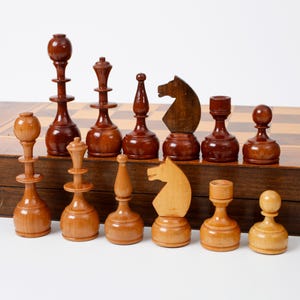 Könnte beinhalten: Ein Set aus hölzernen Schachfiguren in Dunkelbraun und Hellbraun, bestehend aus Königen, Damen, Läufern, Springern, Türmen und Bauern, angeordnet in zwei Reihen. Die Figuren stehen vor einem hölzernen Schachbrett, das als Aufbewahrungsbox dient.