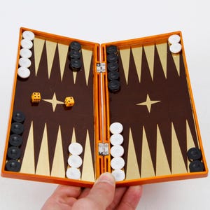 May include: Open backgammon board with brown and gold playing surface, orange frame, and black and white game pieces. Two dice show a four and a one. A classic board game.