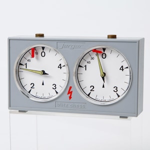 May include: A gray Jerger blitz chess clock with two circular clock faces. Each clock face has black numerals and hands against a white background. The clock is labeled "blitz chess" with a lightning bolt symbol.