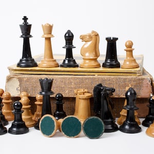 Op de afbeelding: Een verzameling schaakstukken van zwart en licht hout, waaronder koningen, koninginnen, lopers, paarden, torens en pionnen, opgesteld voor een versleten, getextureerde kartonnen doos. Verschillende stukken liggen ondersteboven met groene viltvoetjes.