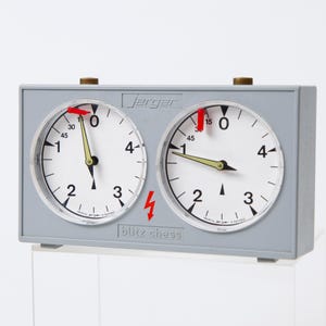 May include: A gray Jerger blitz chess clock with two white clock faces. Each clock face has black numbers and hands, with red indicators at the top. The text "blitz chess" is below the clock faces.