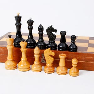 May include: A wooden chessboard with black and white squares. The board is closed and has a set of chess pieces on top. The pieces are made of wood and are painted black and white. There are 12 pieces in total, including a king, queen, bishop, knight, rook, and pawn.