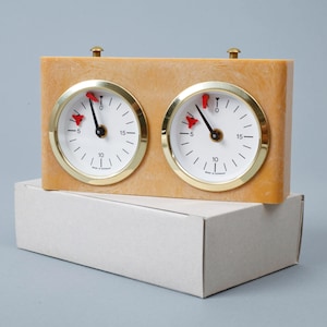 May include: A rectangular analog game clock with a marbled tan casing and two circular white faces featuring black numerals, gold-toned bezels, and red indicator needles. Two brass-colored buttons are positioned on the top edge of the unit.