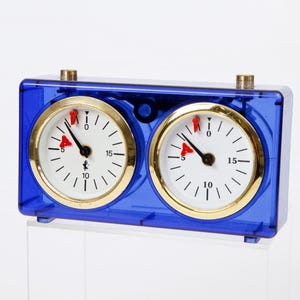 May include: A blue chess clock with two circular white clock faces. Each clock face has gold-colored trim, black hands, and red indicators. The clock is made of transparent blue plastic and has two gold-colored knobs on top.