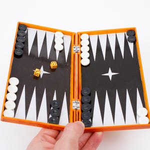 Könnte beinhalten: Ein tragbares Backgammon-Spiel mit einem orangefarbenen Kunststoffrahmen und einer schwarzen Spielfläche mit weißen dreieckigen Feldern. Das Set enthält weiße und schwarze runde Spielsteine auf dem Brett sowie zwei kleine gelbe Würfel.