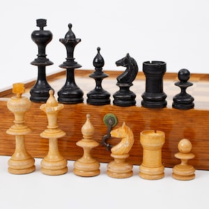 Puede incluir: Un juego de ajedrez completo con piezas negras y crema, que incluye un rey, una reina, una torre, un caballo, un alfil y un peón. Las piezas están dispuestas sobre un tablero de ajedrez de madera, el juego descansa sobre una caja de madera.