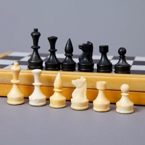 Puede incluir: Un juego de ajedrez con piezas blancas y negras sobre un tablero de ajedrez de madera. Las piezas son de plástico y de estilo clásico. El tablero de ajedrez está cerrado y tiene un marco de madera.