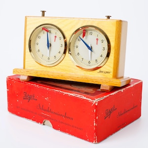 May include: A light wood analog chess clock with two circular white faces, gold-toned bezels, and blue and red hands. The clock features the brand name Jerger on the front and sits atop a rectangular red cardboard box with German text.