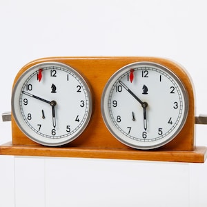May include: A wooden chess clock with two white clock faces, each with black numerals and hands. The clock has a light brown wooden frame and a silver metal trim around the clock faces. The chess piece symbol is above the 6.