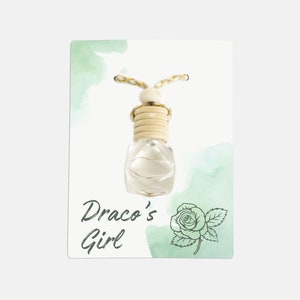 Puede incluir: Una tarjeta blanca con un fondo de acuarela verde presenta una botella de vidrio transparente con un tapón de madera y una cuerda atada. La botella está colgada de la tarjeta. El texto "Draco's Girl" está impreso en la tarjeta en cursiva negra.