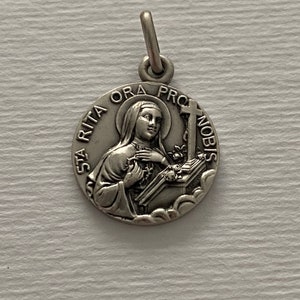 Può includere: Pendente in argento con una raffigurazione di Santa Rita da Cascia, che tiene una croce e un libro. Il pendente è inciso con "Sta Rita Ora Pro Nobis".