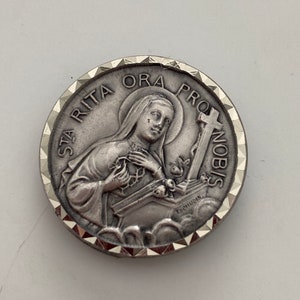 Peut inclure: Médaille religieuse en métal argenté avec une image en relief de sainte Rita de Cascia tenant un cœur et une croix. La médaille a un bord festonné et l'inscription "Sta Rita Ora Pro Nobis".