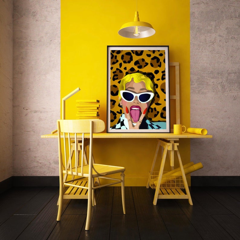 May include: A vibrant yellow home office setup featuring a desk, chair, and a framed pop art print. The artwork depicts a woman with sunglasses and her tongue out, set against a leopard print background. A yellow lamp, books, and accessories complete the stylish workspace.