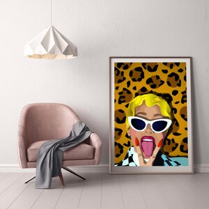 May include: A framed pop art print featuring a woman with blonde hair, white sunglasses, and a leopard print background. The woman is sticking out her tongue and making a peace sign. The print is displayed in a room with a pink chair and a gray blanket.