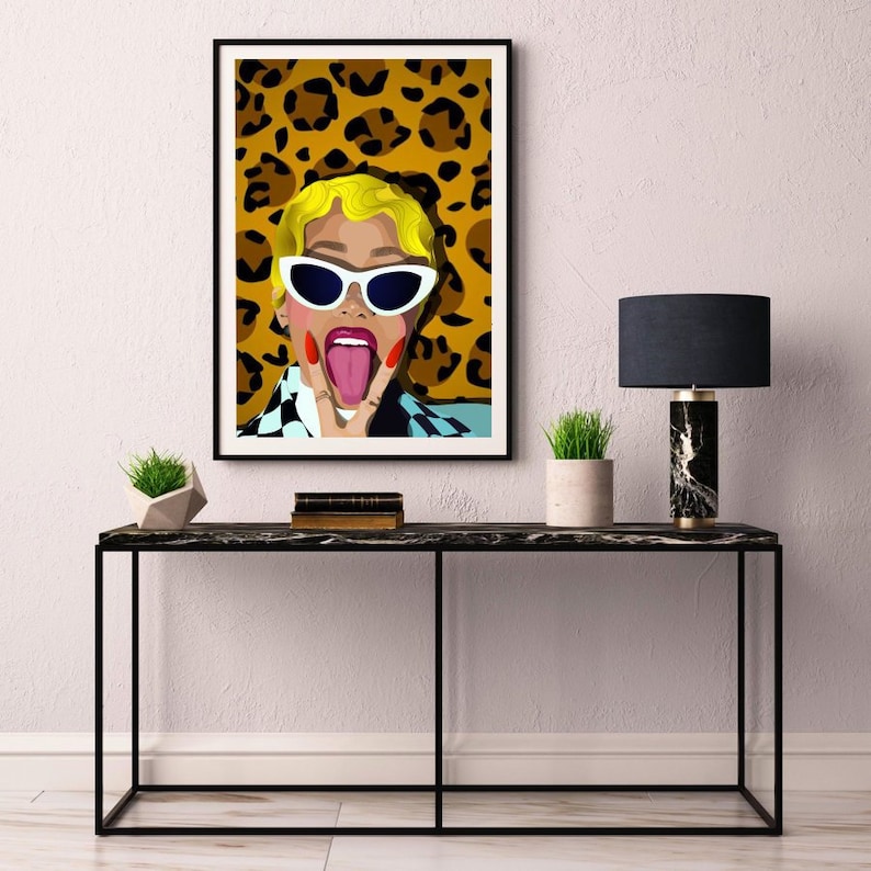 May include: A framed art print featuring a pop art style illustration of a person with blonde hair, white sunglasses, and a checkered shirt. The background is a leopard print pattern. The person is sticking out their tongue and making a peace sign.