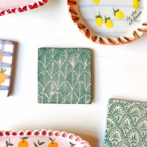 Peut inclure: Un dessous de verre en céramique vert avec un motif géométrique blanc. Le motif ressemble à des feuilles ou des écailles qui se chevauchent.