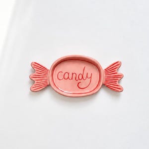 May include: A pink ceramic dish shaped like a wrapped sweet. The centre is oval with the word candy written in red script. The two ends feature red striped detailing. The dish has a smooth, glossy finish and a raised border around the centre.