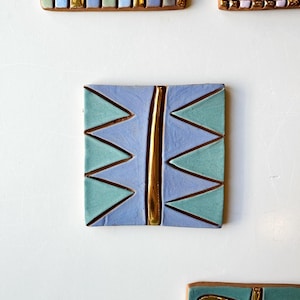 Puede incluir: Un azulejo de cerámica cuadrado con un diseño geométrico. El azulejo está pintado en tonos de azul y verde con una franja dorada en el centro. El azulejo es perfecto para agregar un toque de estilo moderno a cualquier espacio.