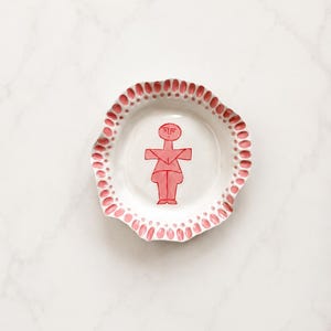 Puede incluir: Un plato de cerámica redondo con un borde irregular y ondulado que presenta un patrón repetitivo de formas rosas similares a pétalos. El centro del plato blanco muestra una figura rosa estilizada y simétrica con cabeza circular.
