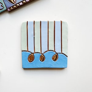 Op de afbeelding: Vierkante keramische tegel met een lichtblauw en groen gestreept patroon, geaccentueerd met goudkleurige druppelvormen en een blauw golfontwerp aan de onderkant. Een andere tegel met een vergelijkbaar ontwerp is gedeeltelijk zichtbaar.