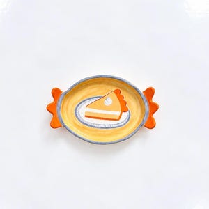 May include: An oval ceramic dish with orange scalloped handles on each side. The interior features a yellow background with a painted illustration of a single slice of pumpkin pie on a small white plate, topped with a dollop of white cream.
