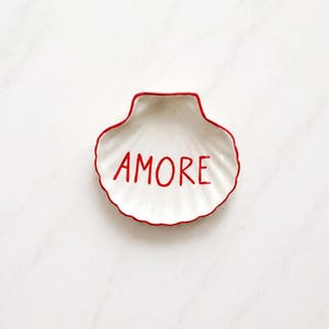 May include: A white ceramic shell-shaped trinket dish with a scalloped edge outlined in red. The word AMORE is written in the center in red, hand-painted style lettering. The dish has a glossy finish and a concave, ribbed interior surface.