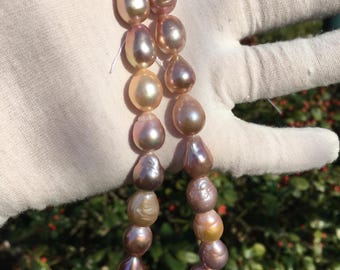May include: A strand of iridescent pearls in shades of pink, peach, and gold. The pearls are strung on a white thread and are displayed on a white gloved hand.