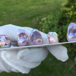 May include: Five iridescent, baroque pearls displayed on a white surface. The pearls have a mix of pink, purple, and gold hues, with irregular shapes. The background is blurred green foliage.