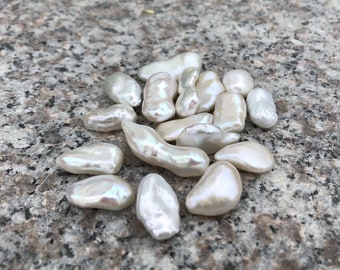 May include: A collection of white baroque pearls, with an irregular shape and a smooth, lustrous surface. The pearls are scattered on a gray, textured surface.