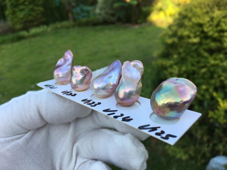 May include: Five iridescent baroque pearls in shades of pink, purple, and green are displayed on a white card. The pearls have irregular shapes and a lustrous surface. The card has black numbers and letters.