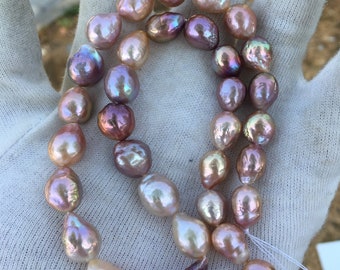 May include: A strand of iridescent baroque pearls in shades of pink, lavender, and white. The pearls are strung on a white cord and are displayed on a white surface.