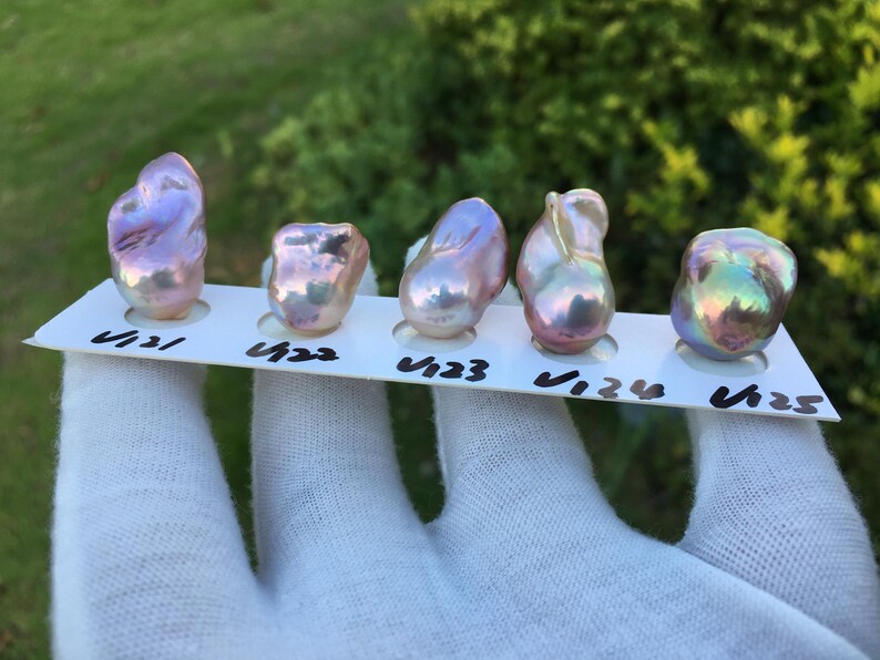 May include: Five iridescent baroque pearls displayed on a white card, each with a unique shape and color. The pearls range in hues from pink and lavender to green and gold. The card has black handwritten numbers.