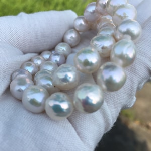 May include: A strand of white pearls, with a slight iridescent sheen, is draped over a white cloth. The pearls are round and smooth, and they are arranged in a loose, flowing pattern.