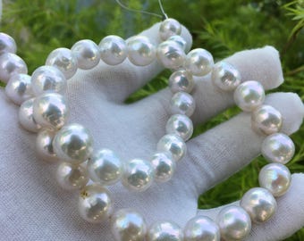 May include: Two strands of lustrous, white pearls. The pearls are round and have a subtle iridescent sheen. The strands are held against a white fabric background, with green foliage visible in the background.
