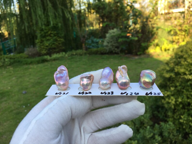 May include: Five iridescent pearls of varying shapes and sizes are displayed on a white card. The pearls exhibit a range of colors, including pink, purple, and gold, with a shimmering, lustrous surface. The card has handwritten numbers.