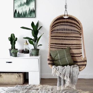May include: A hanging wicker chair with a green cushion and a gray throw blanket. The chair is hanging from a white ceiling with a chain. There is a white dresser with a green plant and a framed print of a forest scene behind the chair.