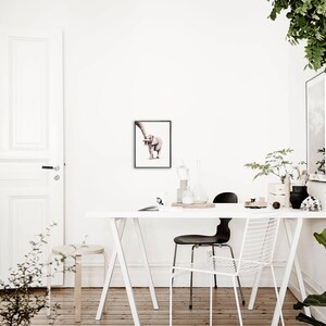 May include: A framed watercolor painting of a baby elephant with its trunk raised, standing on a white background. The painting is hanging on a white wall in a room with a white table and chairs.