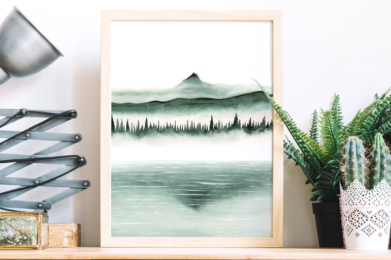 K&ouml;nnte beinhalten: Ein gerahmtes Aquarellbild einer Bergkette mit einem See im Vordergrund. Die Berge sind dunkelgr&uuml;n und von Nebel bedeckt. Der See ist hellblau und spiegelt die Berge wider.