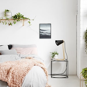 May include: A bedroom interior with a white wall, a bed with a white duvet and a pink knitted throw, a black framed print of a mountain landscape, a black lamp with a gold base on a black side table, and a potted plant on the table.