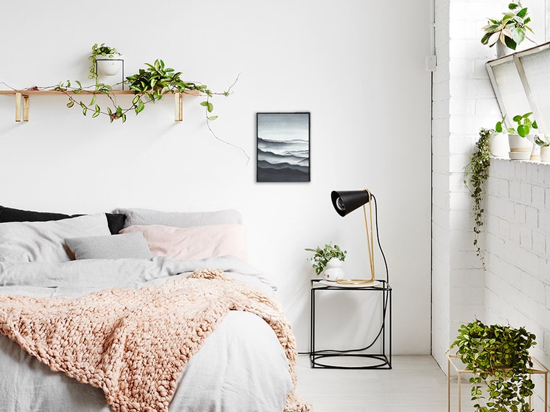 May include: A bedroom interior with a white wall, a bed with a white duvet and a pink knitted throw, a black framed print of a mountain landscape, a black lamp with a gold base on a black side table, and a potted plant on the table.