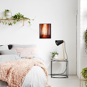 May include: A bedroom interior with a white wall, a bed with a white duvet and a peach-coloured throw blanket, a black side table with a lamp, and a framed painting of a forest path with a golden glow.