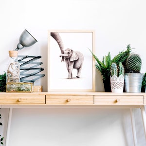 May include: A framed watercolor painting of an elephant and a baby elephant walking together, displayed on a wooden desk with a lamp, a glass bottle, and potted plants.